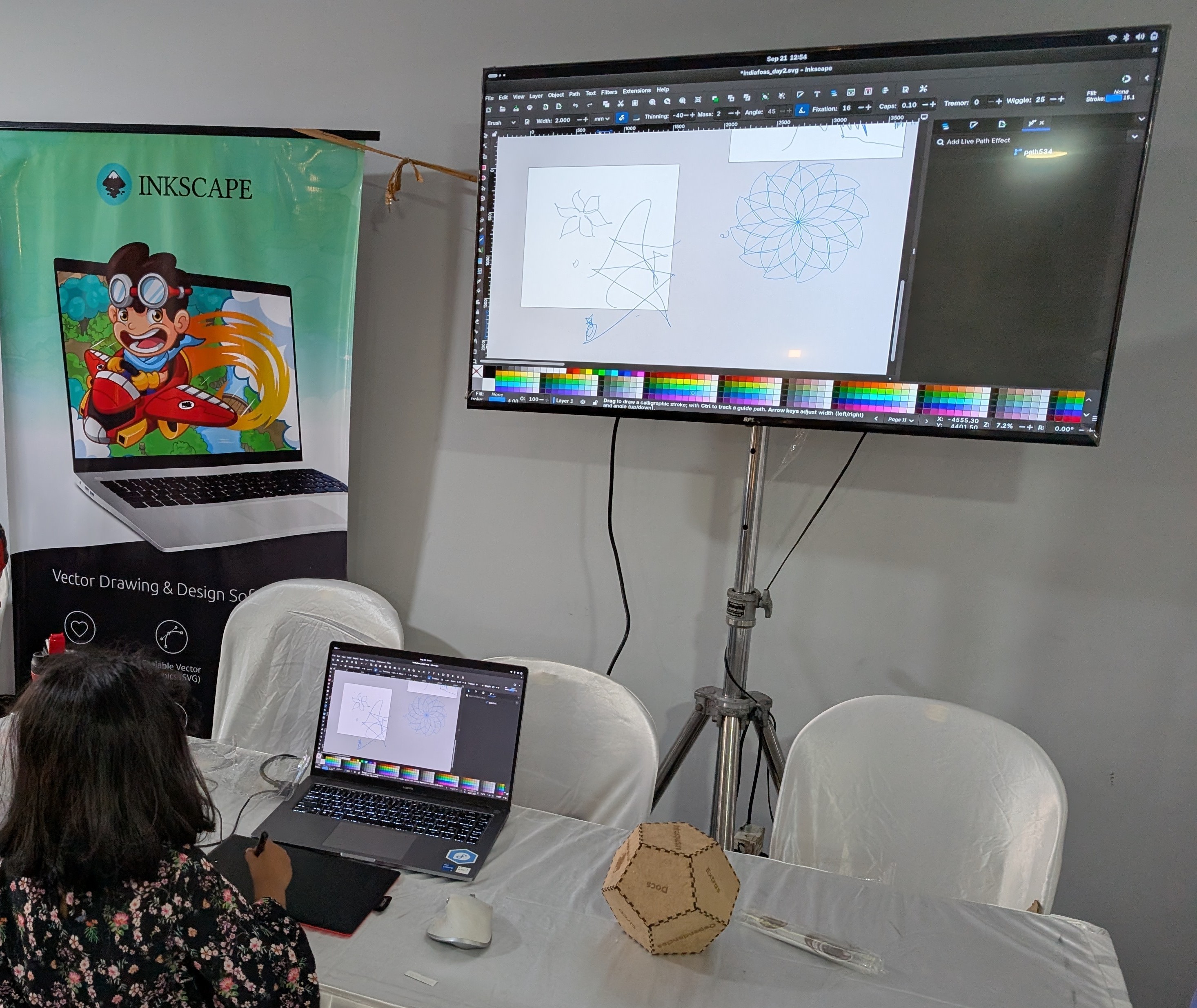 Inkscape booth at IndiaFOSS 2025. The booth has a laptop connected to a mouse and a drawing tablet, running Inkscape 1.4.2. Behind the laptop is an Inkscape standee and a TV screen mirroring the laptop.